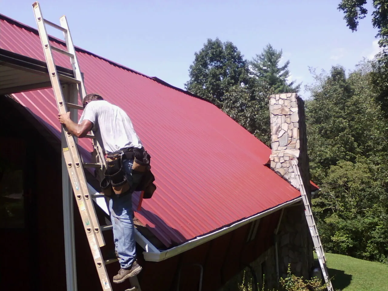Roofer on a ladder beside a red metal roof and stone chimney — Soffit & Fascia Repair by Roofing Brothers in Blissfield